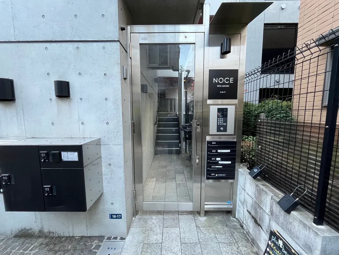 - モダンなアパートのエントランス、透明な扉と金属製の郵便受け、周囲には緑の植栽があり、洗練されたデザインの外観