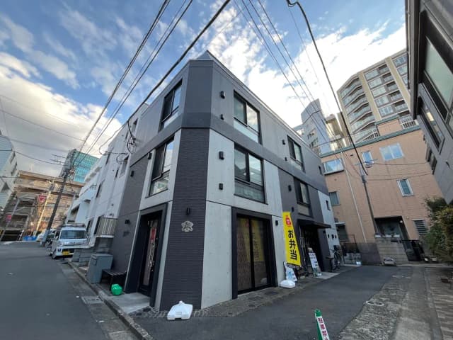 - 現代的な外観の三階建てビル、黒とコンクリートのデザイン、周囲の建物と道路の様子、青空と電線が見える街並み