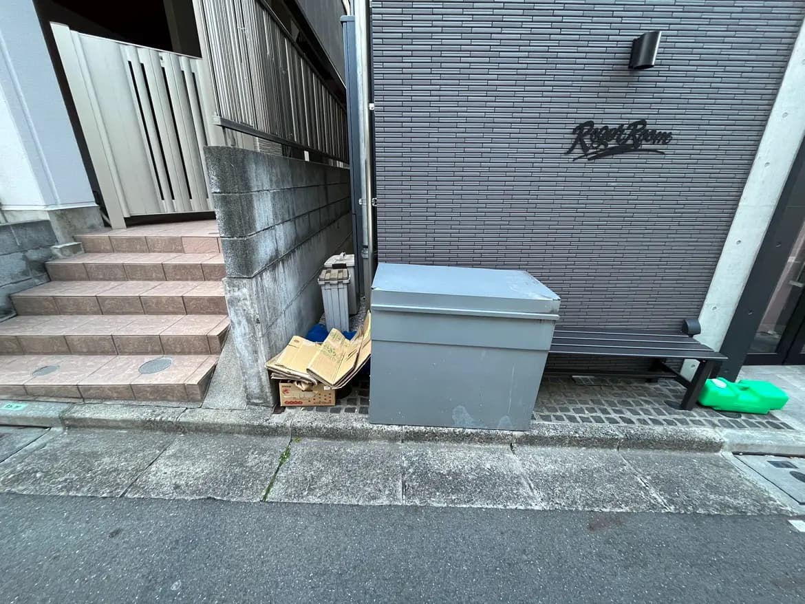 - 鉄筋コンクリートの壁の脇に置かれた大型のごみ箱、近くに段差のあるタイルの階段と木製のベンチ、周辺には雑誌が置かれた段ボール箱が見える街の一角