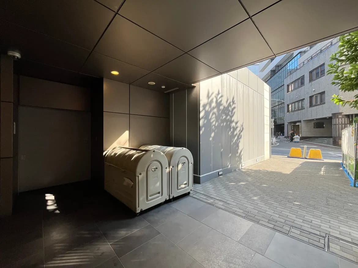 - モダンな建物のエントランス、右側に二つの白いゴミ箱が配置された明るい空間、外には舗装された道と人々の姿が見える