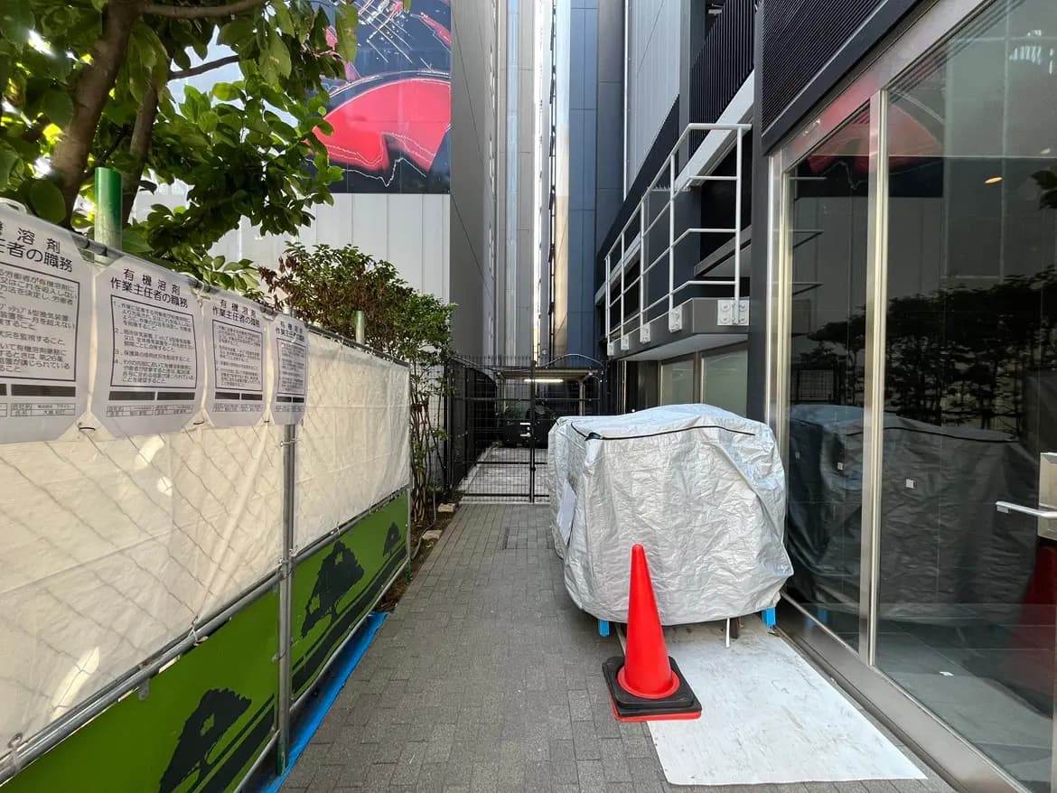 - 建物の間の狭い通路、工事用のフェンスと看板、ビニールシートで覆われた物品、赤いコーンが置かれた現場の様子
