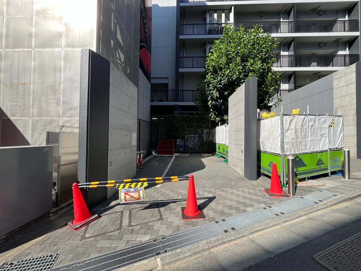 - 建設中の敷地入口、コーンとフェンスで区切られた道、周囲には現場作業用の資材が置かれたエリア