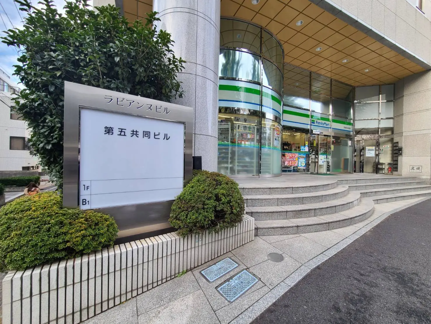 都市のオフィスビル入り口、コンビニエンスストアが地下にあり階段と緑の植栽が見える外観