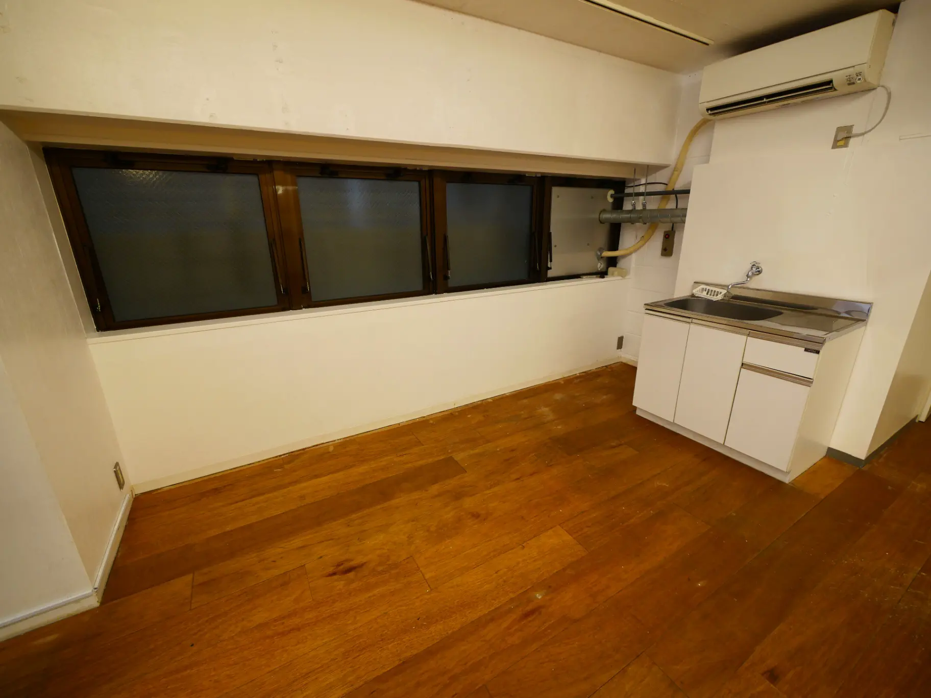 シンプルなキッチン付きのワンルーム、木製フローリング、白い壁、エアコンと小窓が特徴的なインテリア