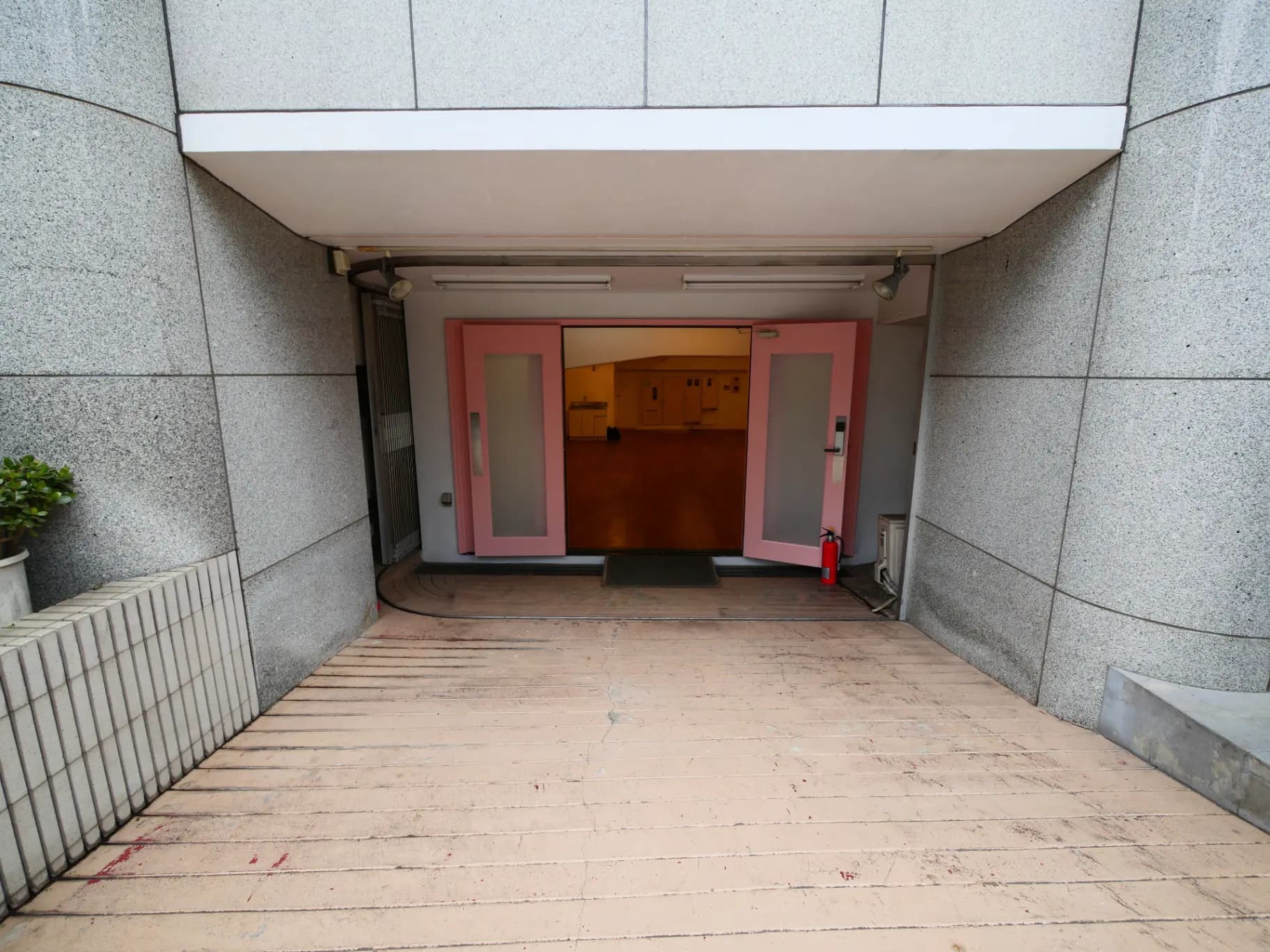 グレーの壁に囲まれた建物の入り口、ピンク色のドア、屋外植物と消火器