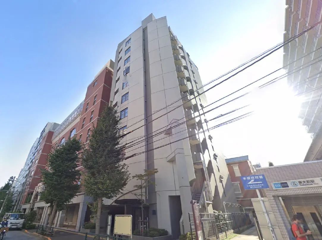 - 都市型マンションの外観、近隣に樹木や電柱があり、晴れた青空が広がる風景