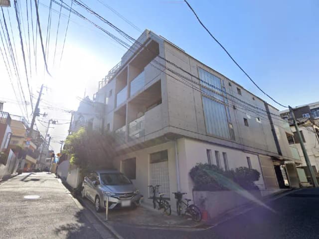 - 日差しを浴びるコンクリート製の現代的なアパートメント、隣に駐車された車と自転車、周囲に電線が張り巡らされた街並み