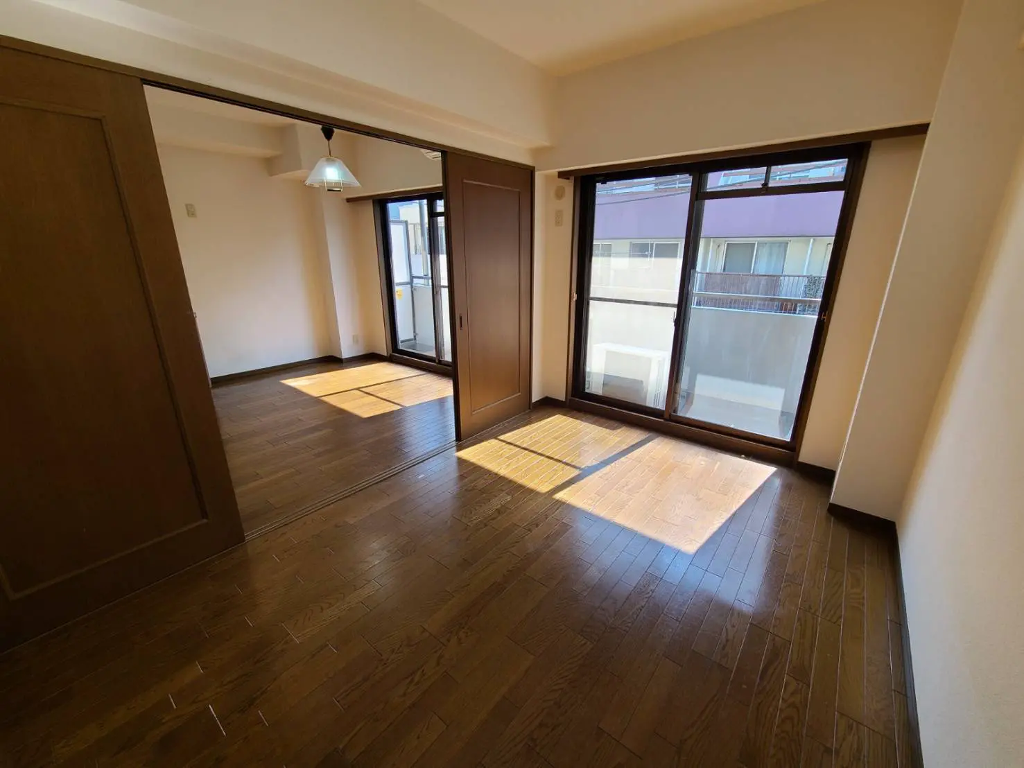 明るい日差しが差し込む空の部屋、ダークブラウンのフローリング、白い壁、バルコニーへと続く大きなガラスの扉