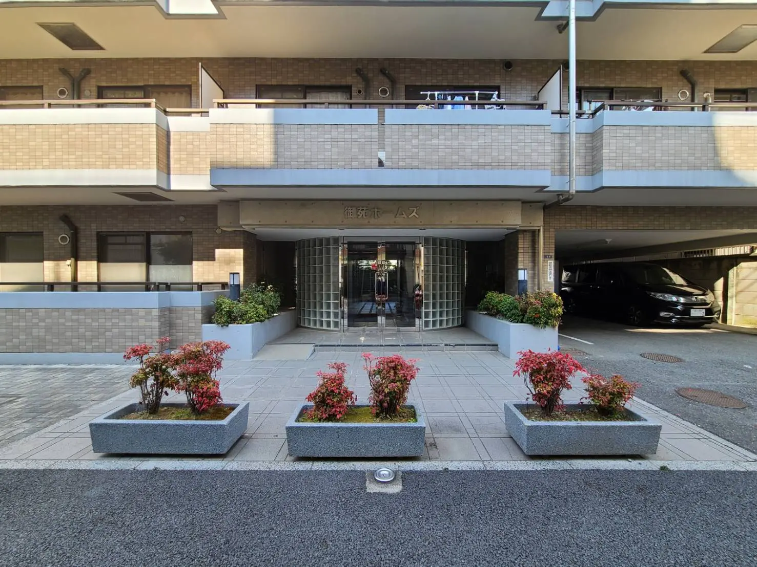 モダンなデザインのマンションエントランス、花壇に赤い植物が植えられ、バルコニーとカーポートがある外観