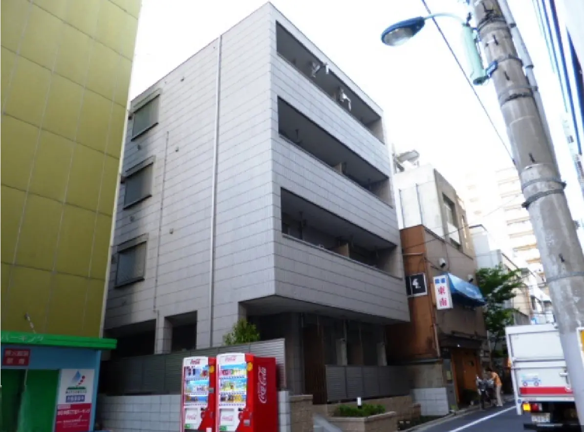 - モダンな外観のアパートメント、銀色の外壁とバルコニー、周囲には飲食店と街灯が立ち並ぶ商業エリア