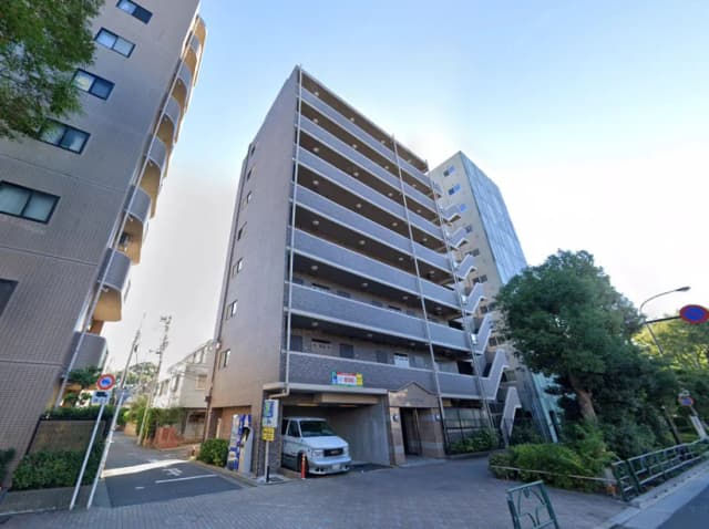 - 都市型マンションの外観、周囲に緑豊かな植栽と近隣の建物が立ち並ぶ風景、明るい青空が広がる景観
