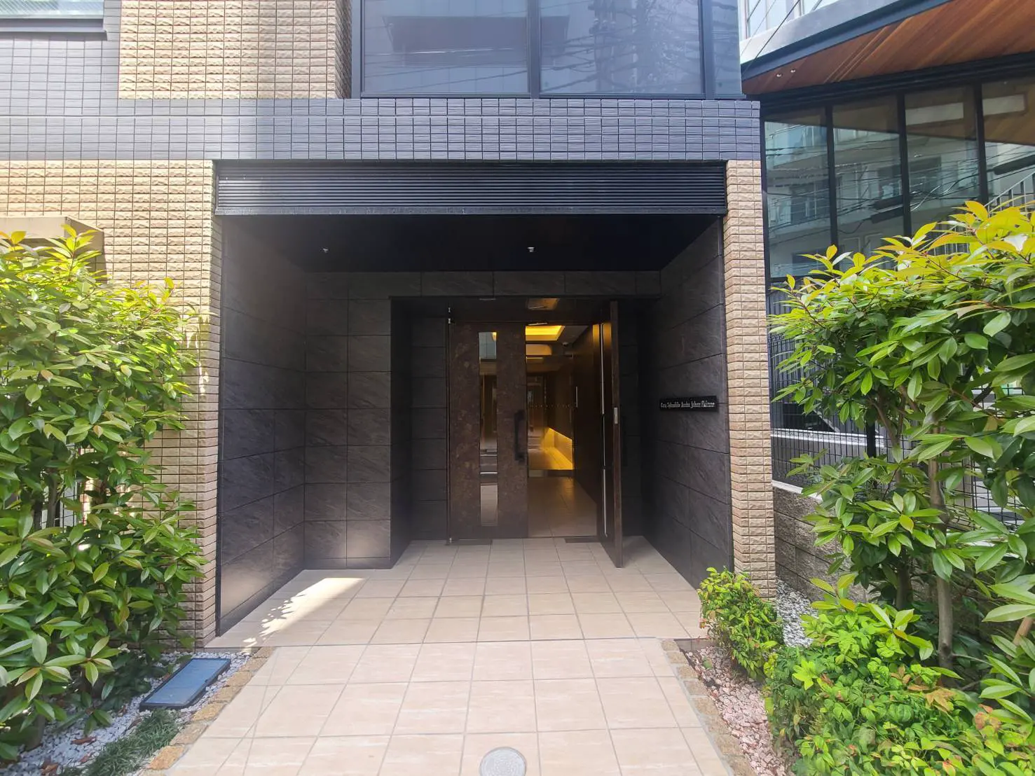 - モダンな外観のマンションエントランス、タイル張りの床と美しい植栽、ガラスドアで明るさを取り入れた入口