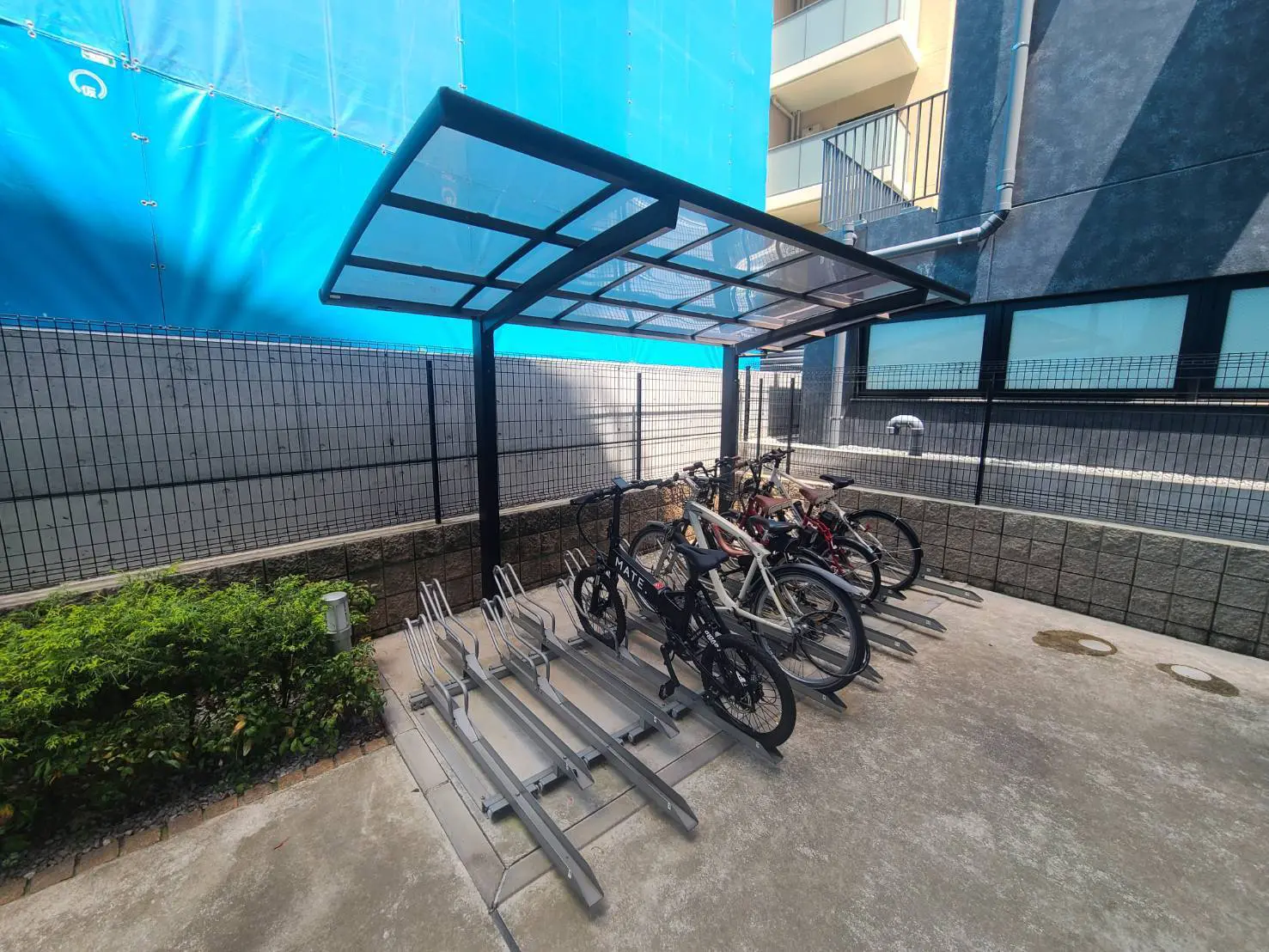 - 自転車が停められた屋根付きの自転車置き場、清潔感のあるコンクリート床と緑の植栽がある外観