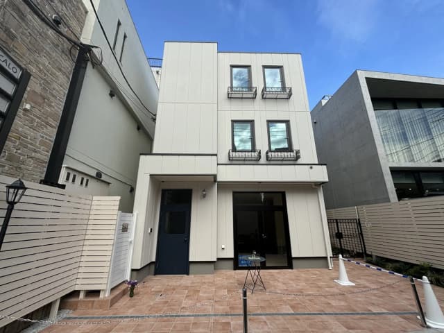 現代的な外観の新築一戸建て、ベージュの外壁と黒い屋根、石造りのアクセント、入口に隣接したブラウンタイルの広場