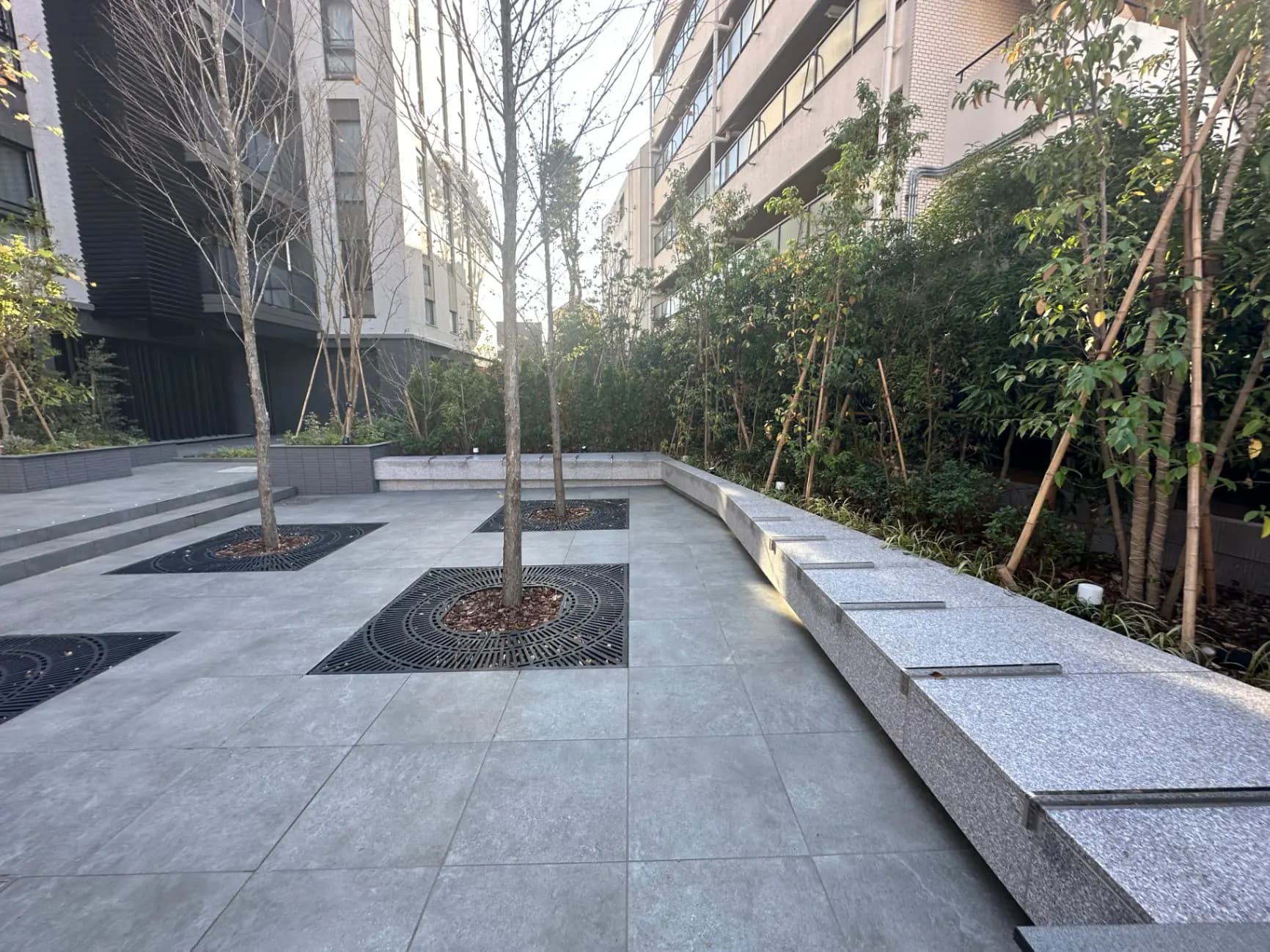 都市型マンションのモダンな共用庭園エリア、グレーのタイルとベンチ、緑豊かな植栽デザイン