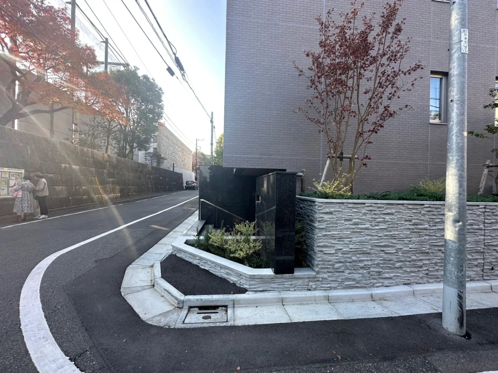 市街地の曲がり角にある石積みの壁と赤く色づく木、道路標識と街灯のある風景