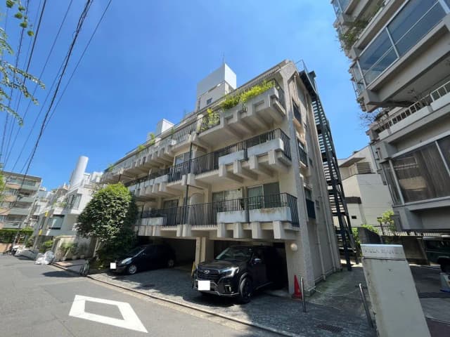 - 明るい青空の下にある3階建てのアパートメントビル、バルコニー付き、周囲には緑の木が点在する住宅街の風景