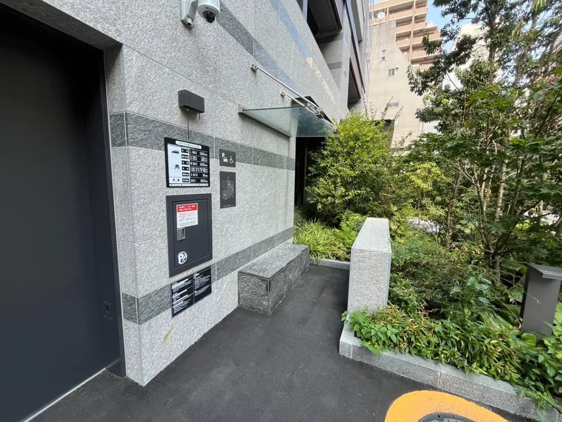 - モダンなビルのエントランス、隣接する緑豊かな植栽と防犯カメラ、案内板が設置された入り口の風景