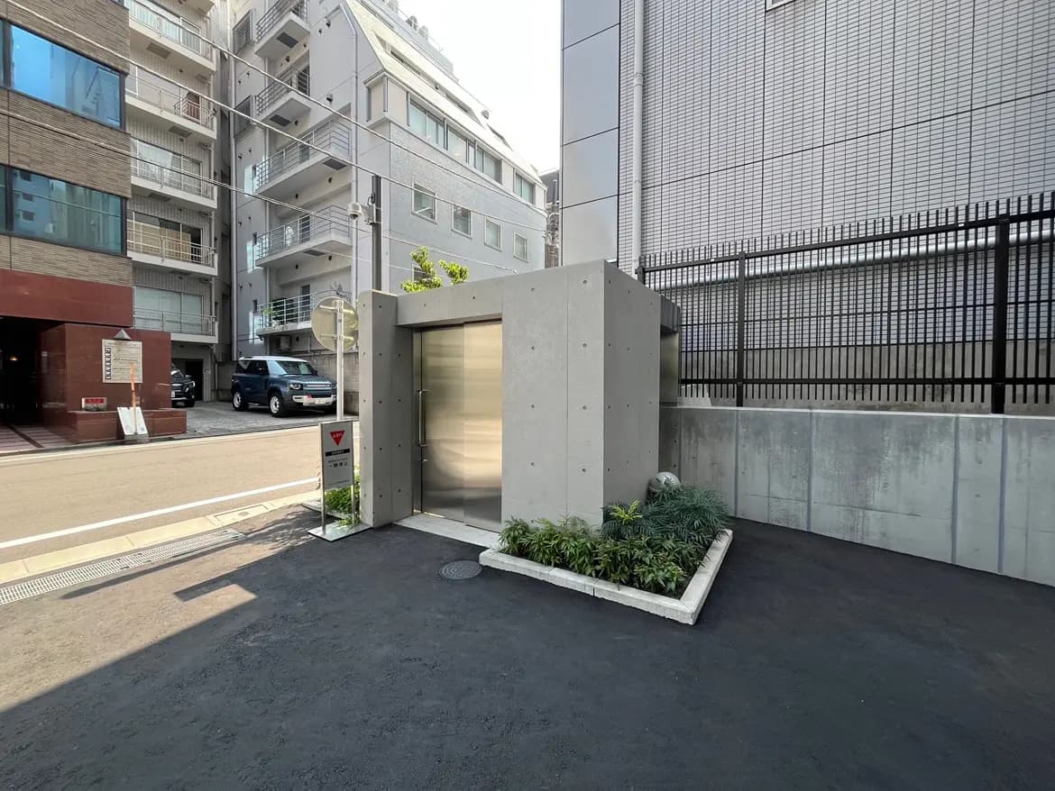- コンクリート造のモダンな外観の入口、周囲に緑の植栽と舗装された道路の風景