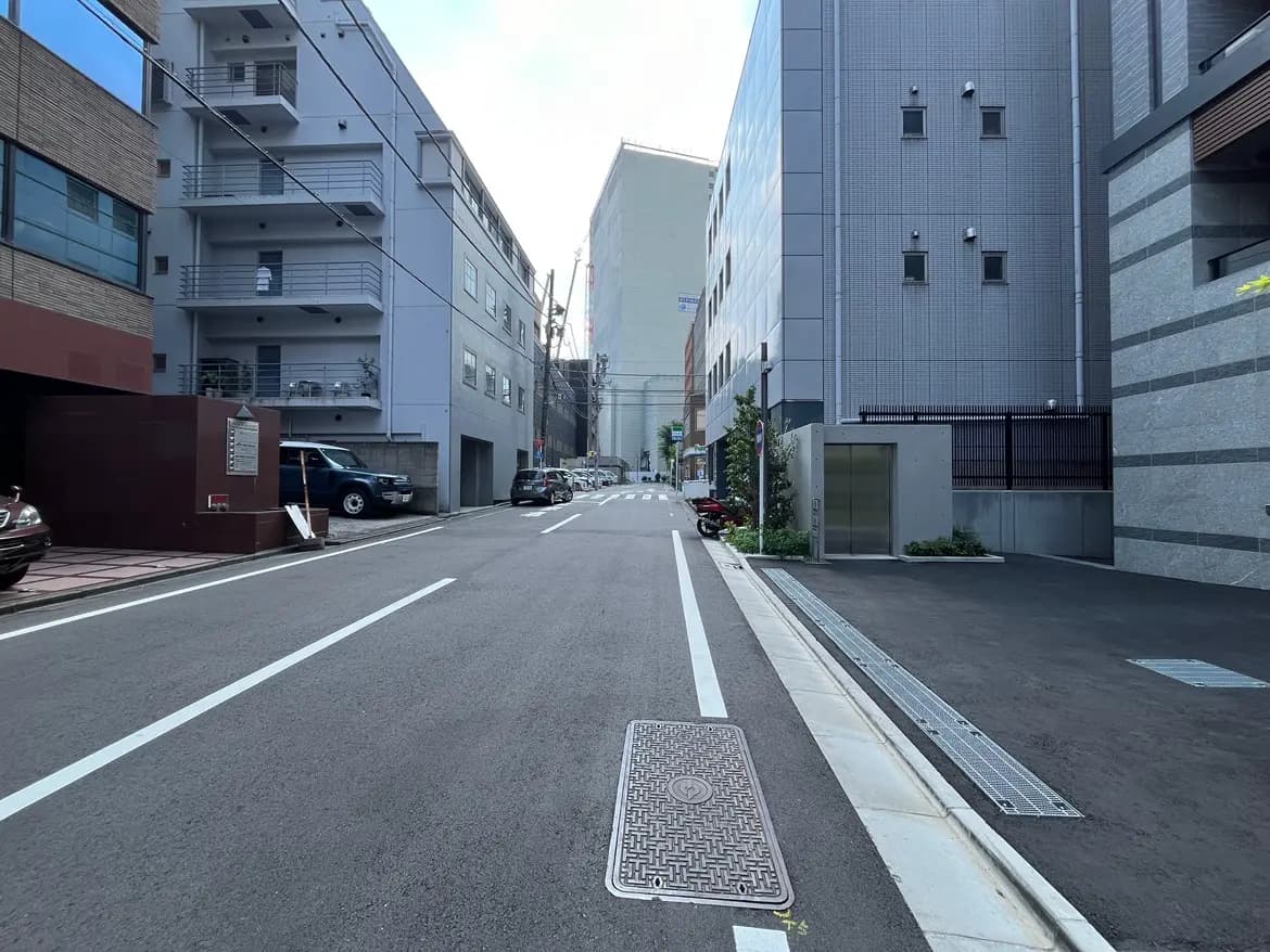 - 都市の静かな通り、両側に立ち並ぶモダンなビルと駐車場、明るい空が広がる風景