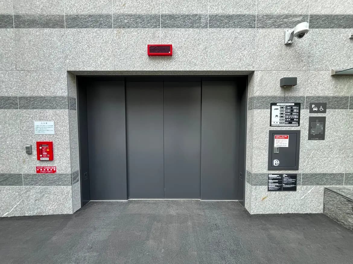 - グレーの石材で作られた建物の外観、黒いエレベーターの扉、周囲には防犯カメラと案内板が設置されているエントランスの風景