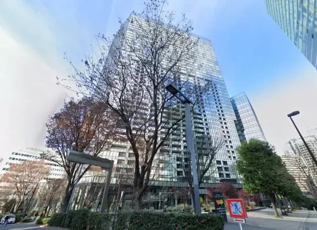 - 高層ビルの外観、周囲に整備された庭木と歩道、青空の下での都市の風景