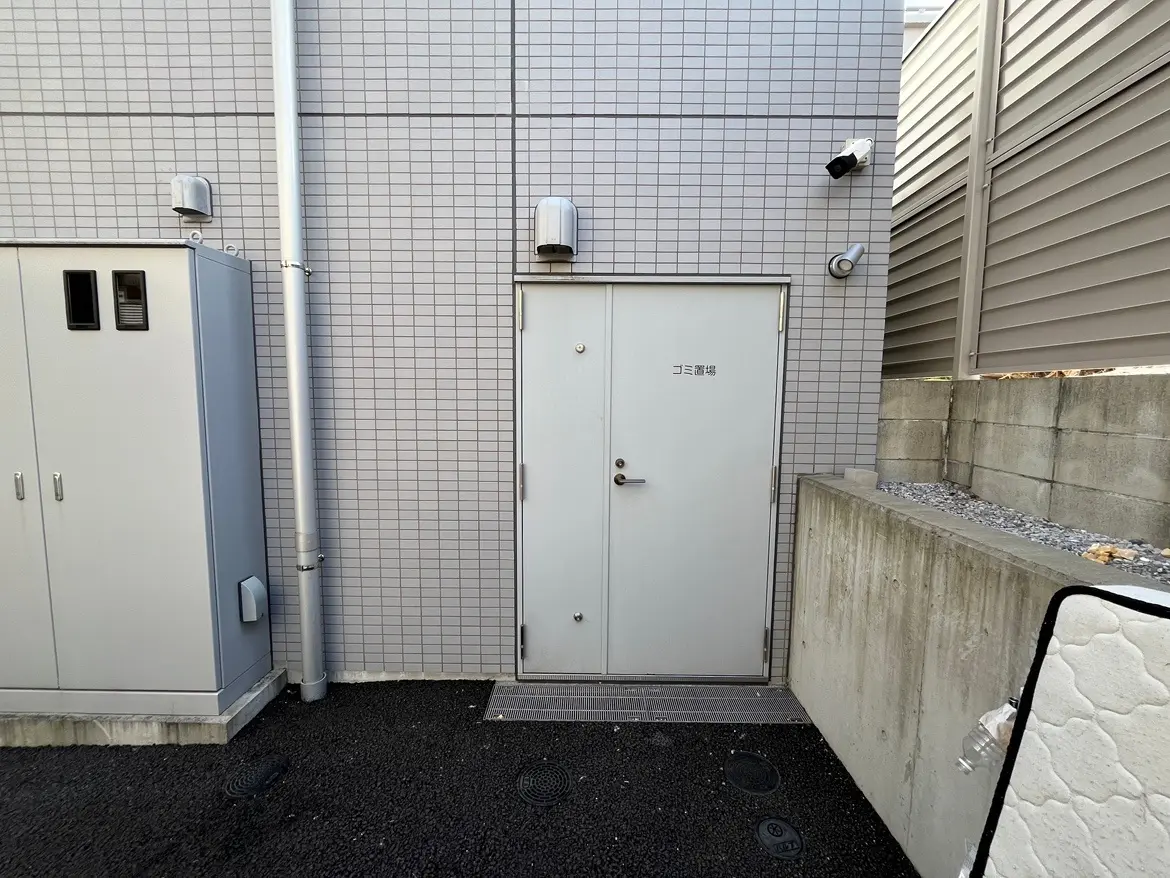 建物の外壁に沿った、ユーティリティエリアの扉と配管、灰色タイルの壁、セキュリティカメラが設置された都市的なシーン