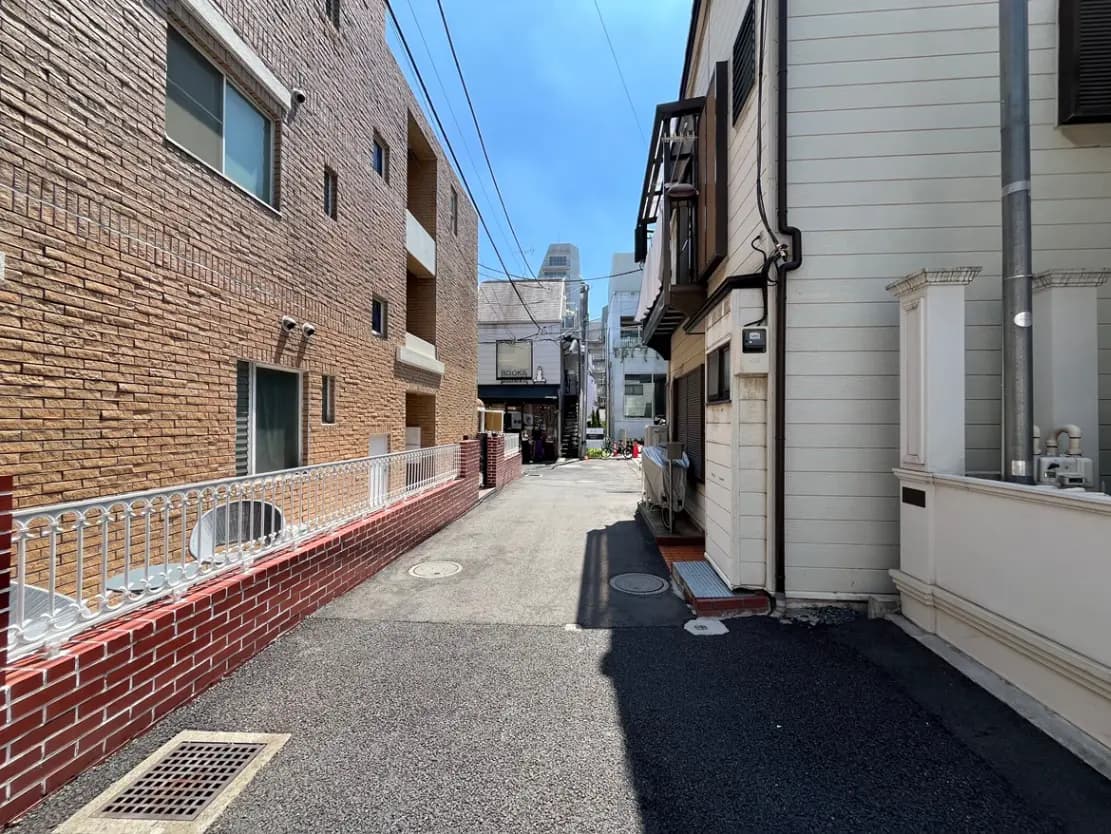 - 日差しの下、レンガの壁と白いフェンスに囲まれた静かな路地、奥に見える建物と青空が特徴的な風景