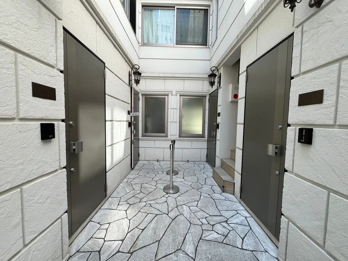 - モダンなデザインのアプローチ、白い石壁と金属製のドア、タイル敷きの床とブラケット照明のエントランスエリア