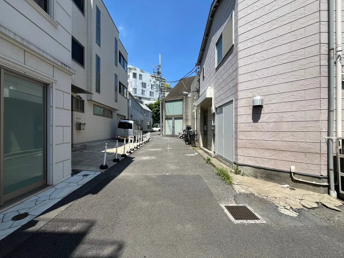 - 明るい青空の下、細い道路に面した現代的な建物と自転車が並ぶエリア、左側には白い壁の建物が見える街の風景