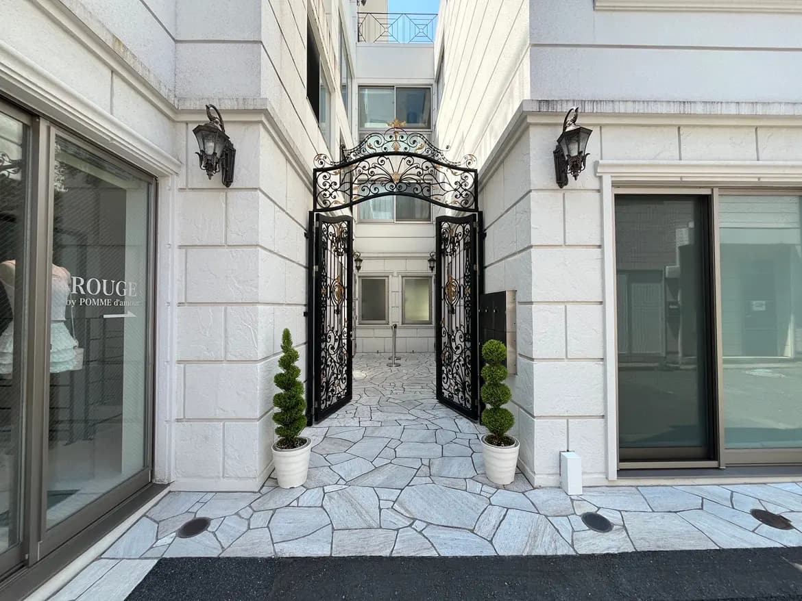 - エレガントなアイアンゲートと石畳のアプローチが特徴的な住宅のエントランス、両側に観葉植物が配置された外観