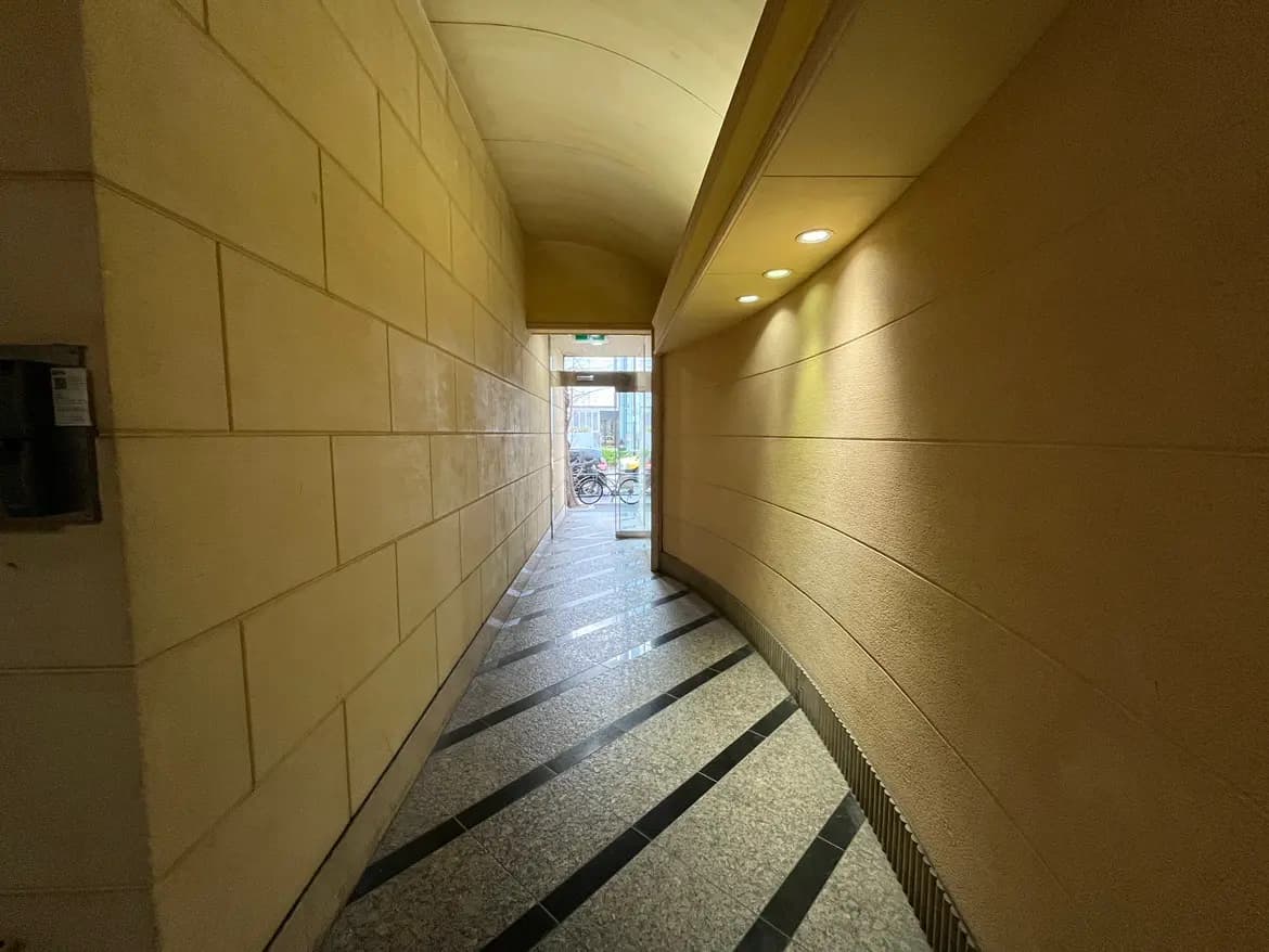 アーチ型の天井と石畳風の床が特徴の照明付きの廊下、通りに面した出口が見える