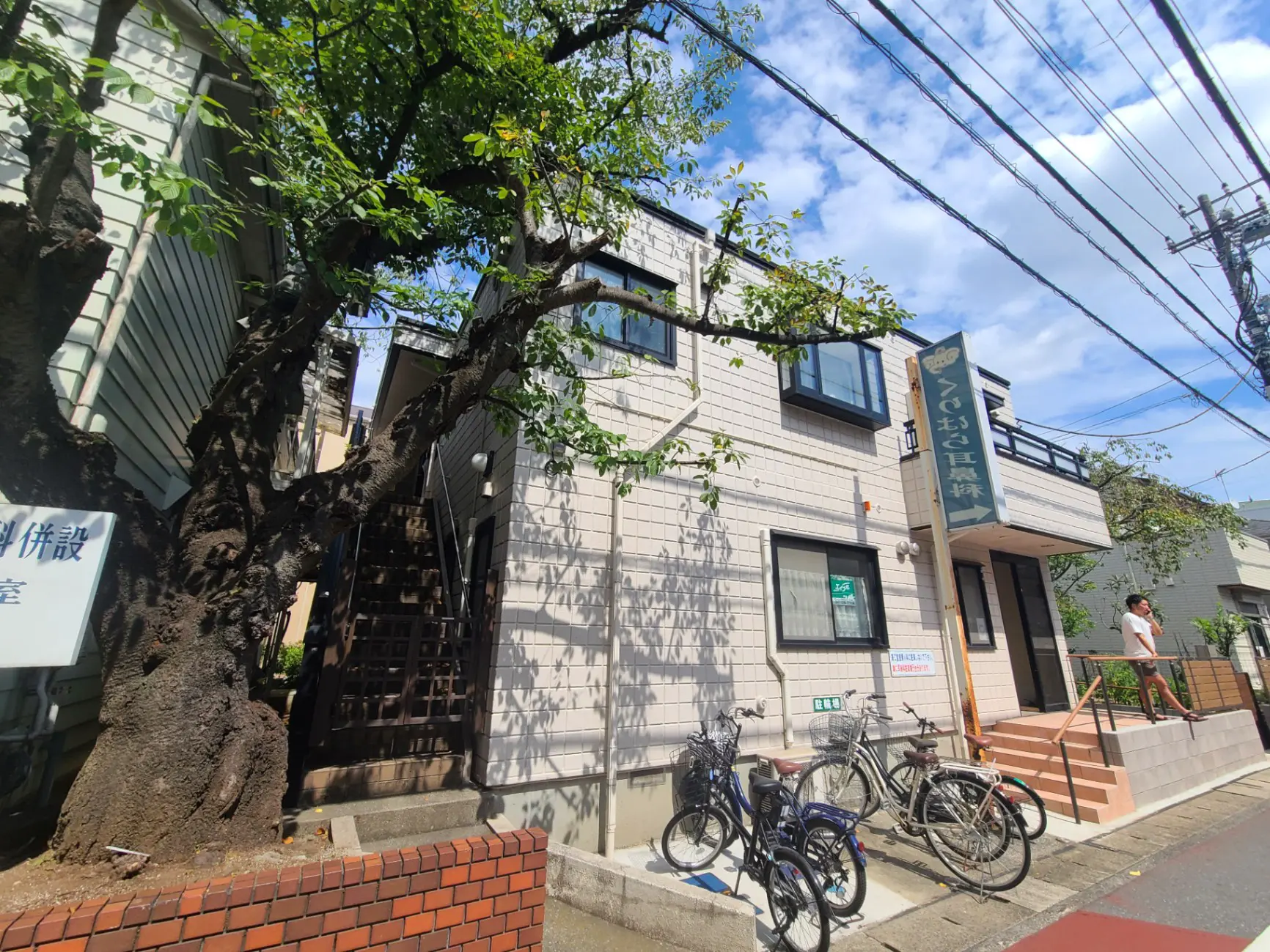 都市の風景、歩道に駐輪された自転車、隣接する建物の影に覆われた大きな木、晴天のもとでの日本のクリニックの外観