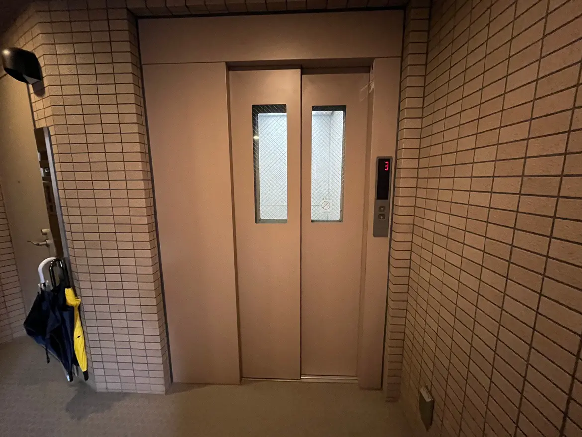 - シンプルなデザインのエレベーターの扉、周囲はレンガ調の壁で囲まれた空間、傘がかけられたハンガーが見えるエントランスの内部