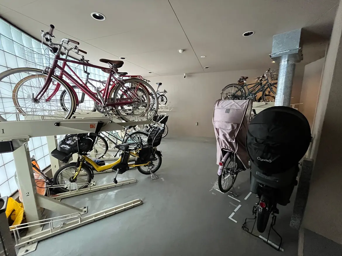 - 自転車が収納された駐輪場の内部、高さのあるラックに複数の自転車が並び、手前にはバイクや子供用自転車も見える風景