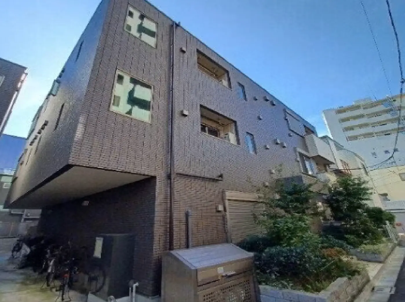 - モダンな外観のアパート、レンガ調の壁、緑豊かな庭と自転車置き場、青空を背景にした住宅街の一角