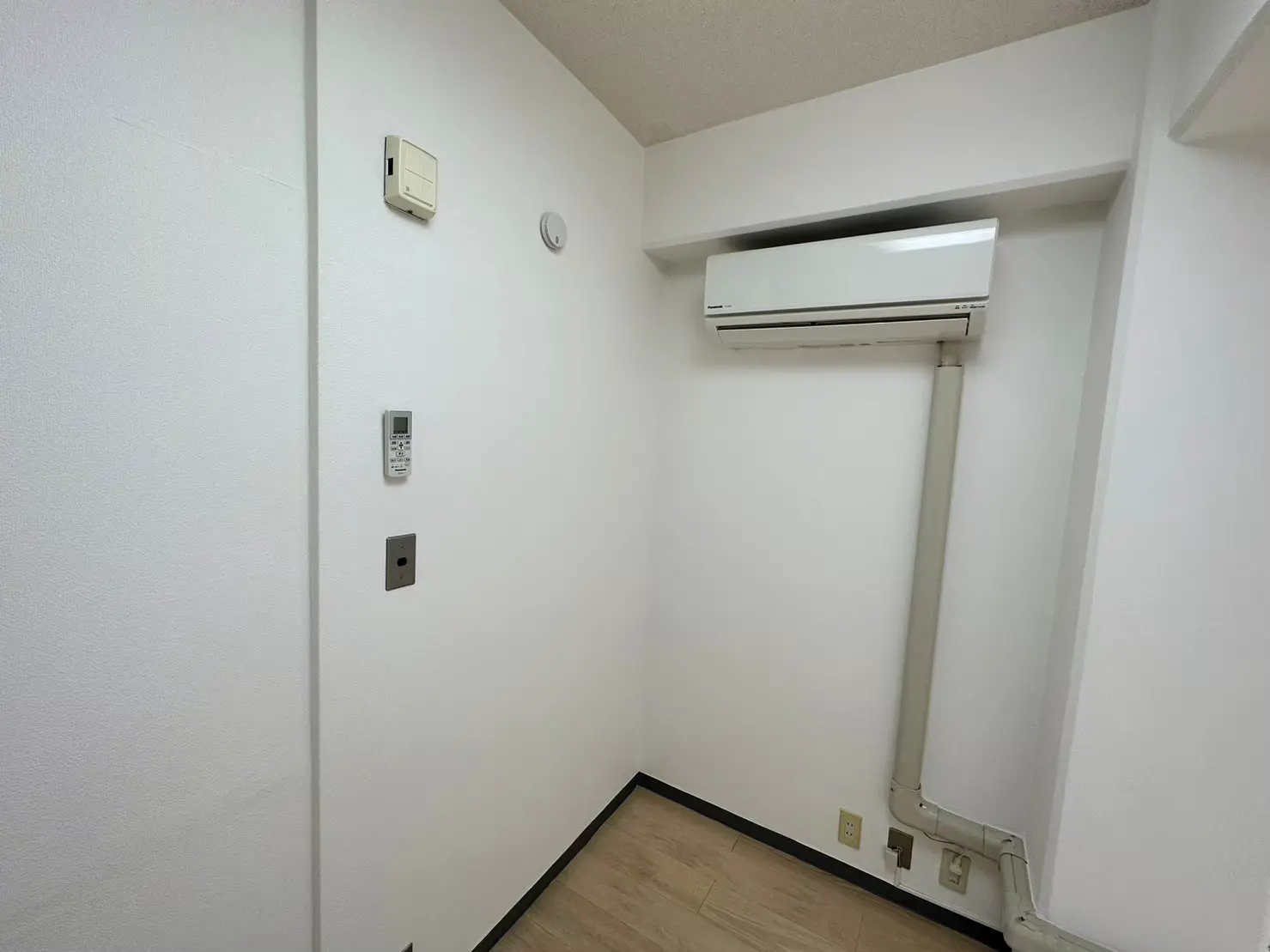 シンプルな白い壁の部屋の隅に設置されたエアコン、壁掛け式温度コントローラーとコンセント