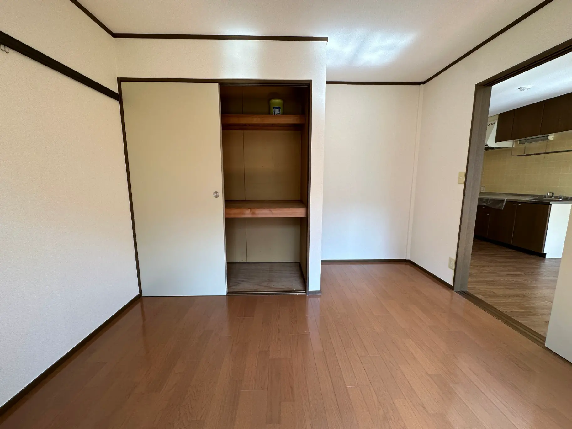 シンプルなデザインの空き部屋、茶色のフローリング、白い壁、クローゼットとキッチンの見えるレイアウト
