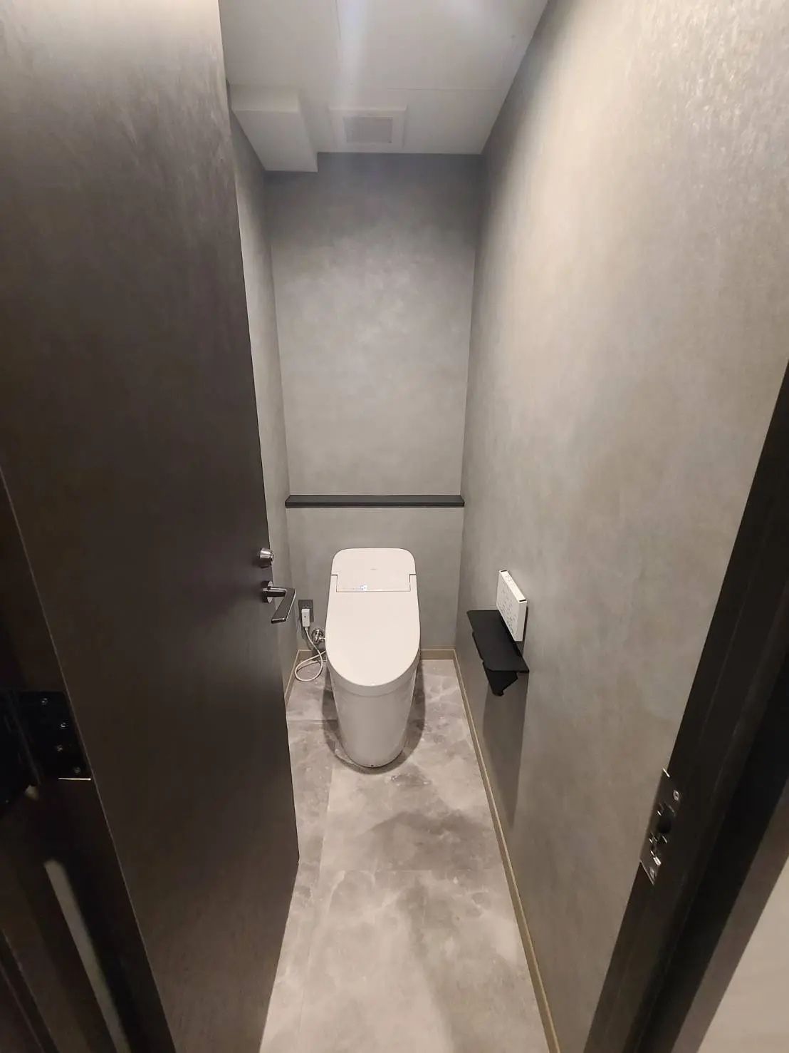 モダンなデザインのトイレ、グレーの壁と床、白い便器と壁掛け式のトイレットペーパーホルダー