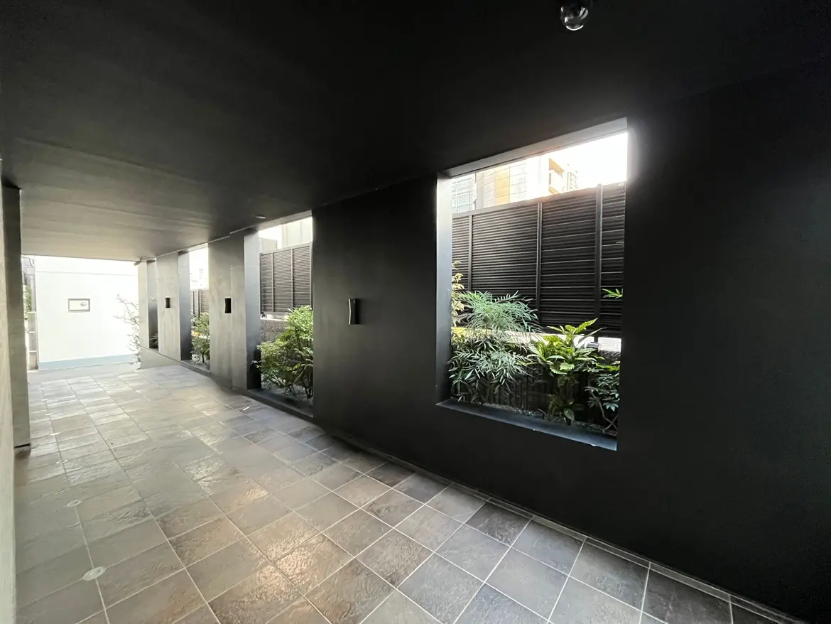 モダンな外観の建物のアプローチ、グレーのタイル張り、壁には緑植物が飾られたニッチがあり、自然光が差し込む空間