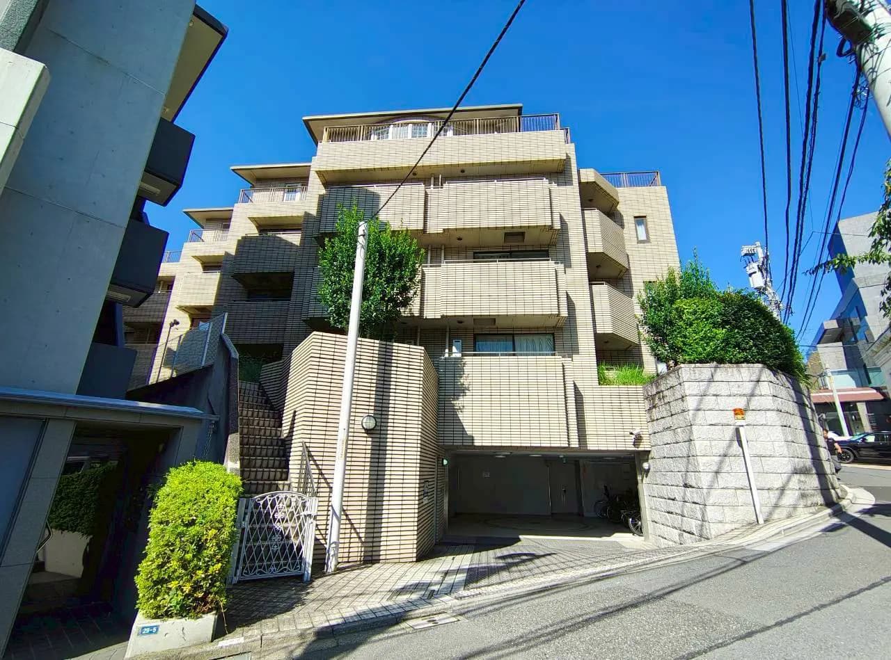 - 明るい青空の下にあるモダンなマンション、前面にはガーデンとアプローチ、周囲には緑豊かな景観が広がる外観