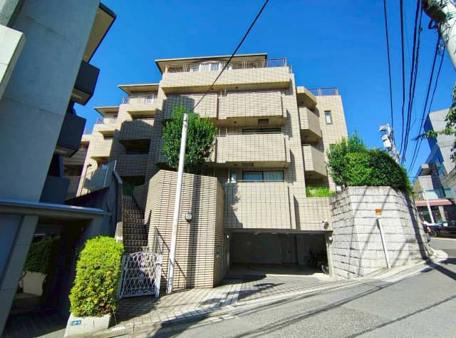 - 明るい青空の下にあるモダンなマンション、前面にはガーデンとアプローチ、周囲には緑豊かな景観が広がる外観