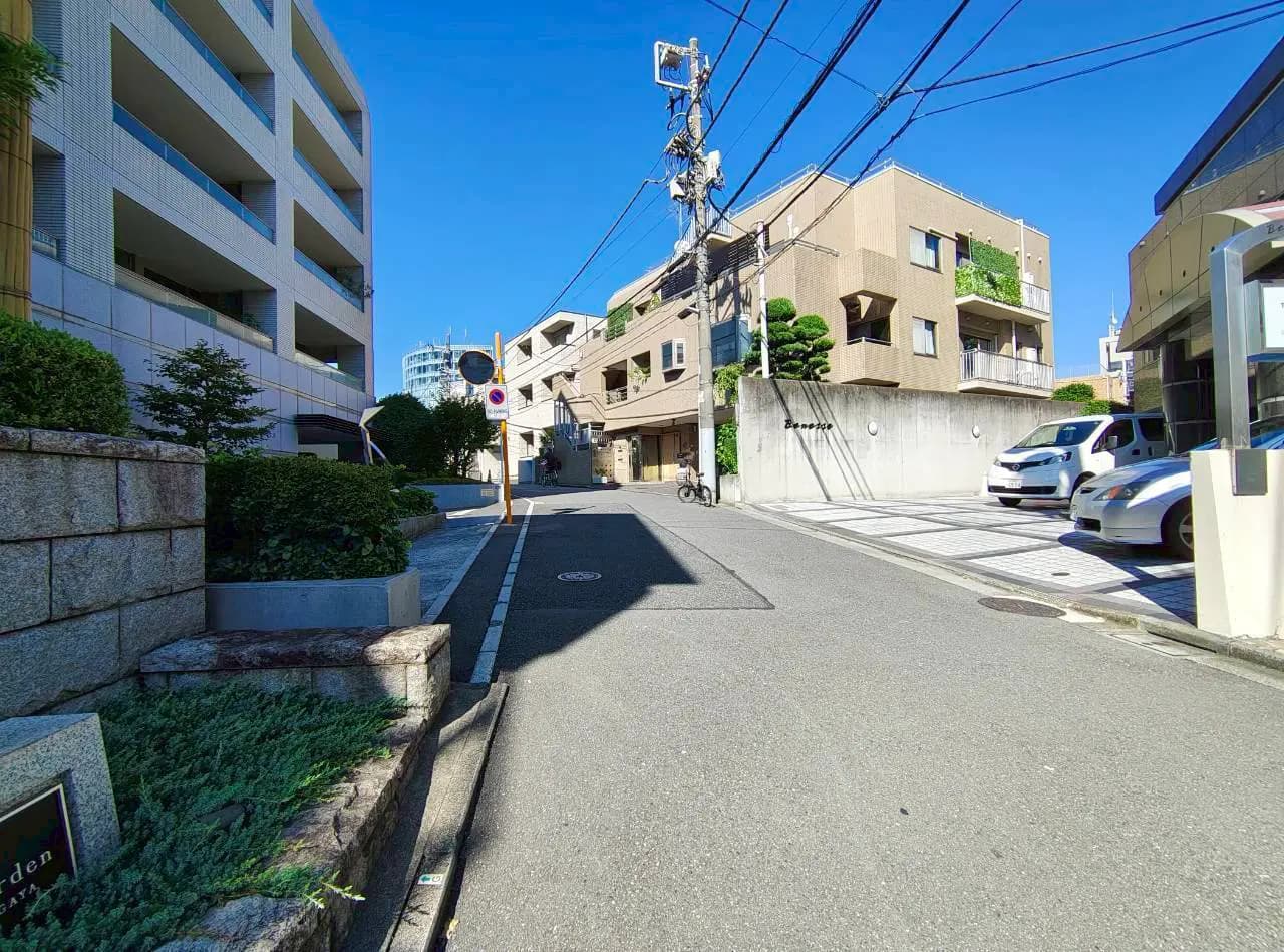 - 青空の下、住宅街の静かな通り、左に並ぶモダンなアパートと右にある車が停まった駐車場、手前に石のガーデンと植物が見える景色