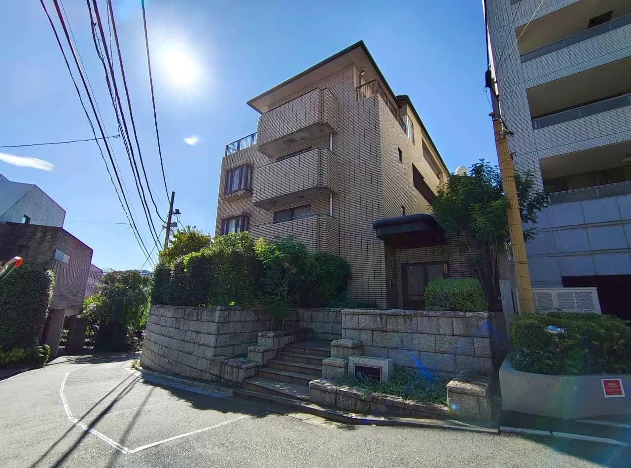 - 明るい空の下に立つ現代的な外観のアパートメント、緑の植栽に囲まれた入り口と石の階段が特徴的な建物