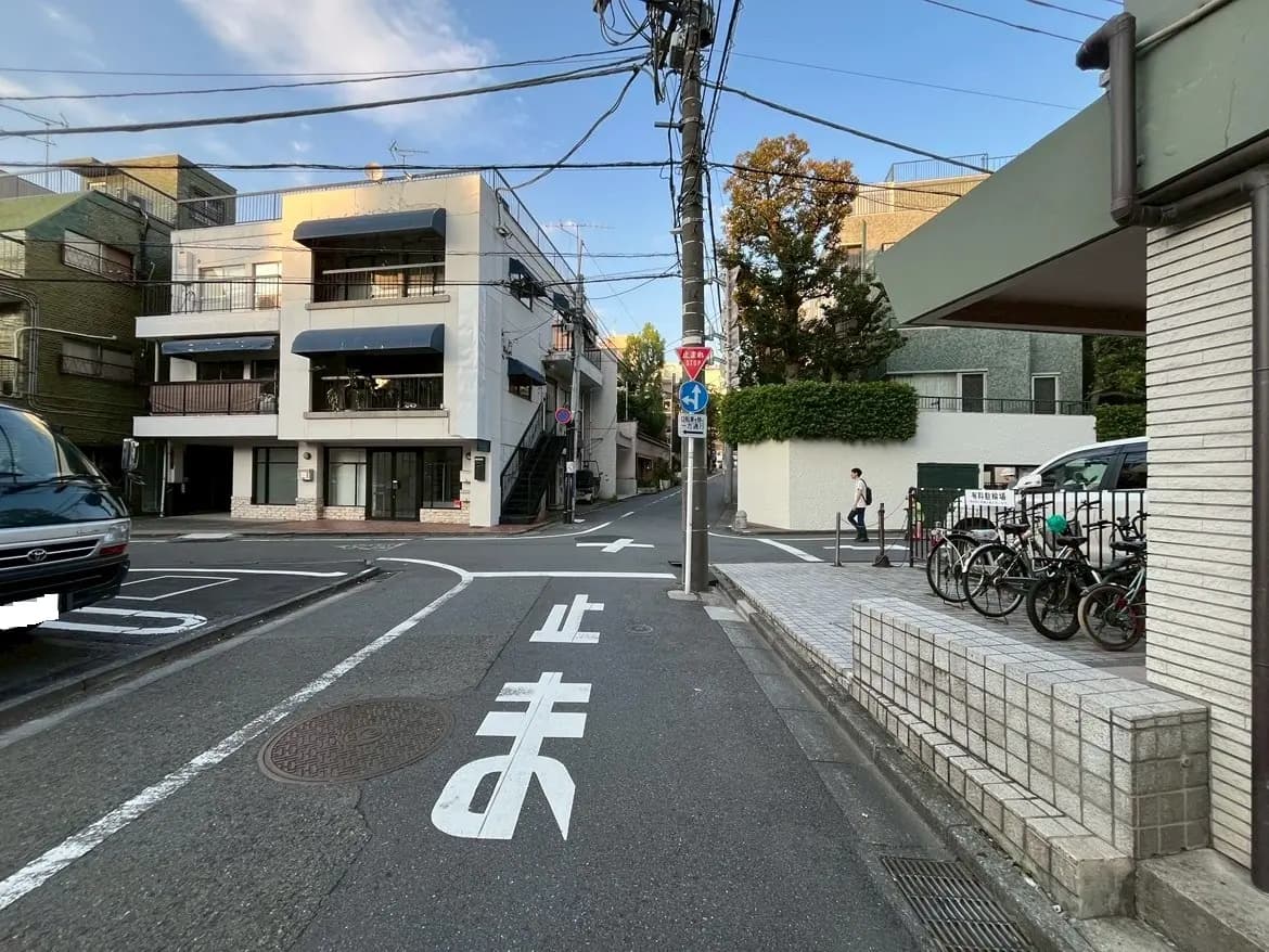 - 都市の風景、住宅街の交差点、左側に商業ビルと車、右側に自転車が停まっている様子、青空と電柱が見える街並み