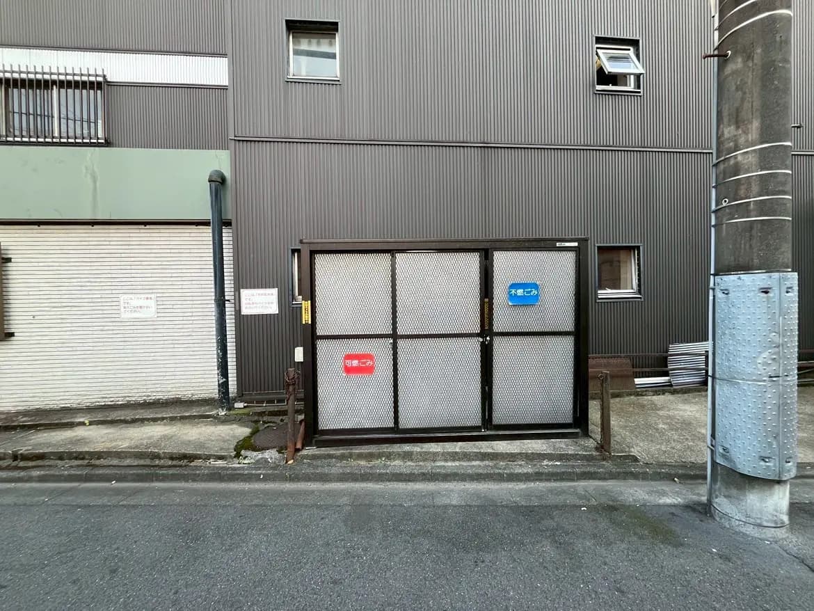 - 鉄製のフェンスとコンクリートの歩道、建物の裏口が見える街の風景、ゴミ捨て場のサイン付き