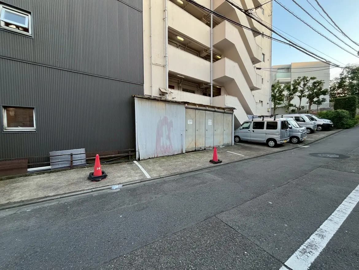 - 駐車場と外壁、近くに停車している複数の車、歩道と赤いコーンが並ぶ通りの風景
