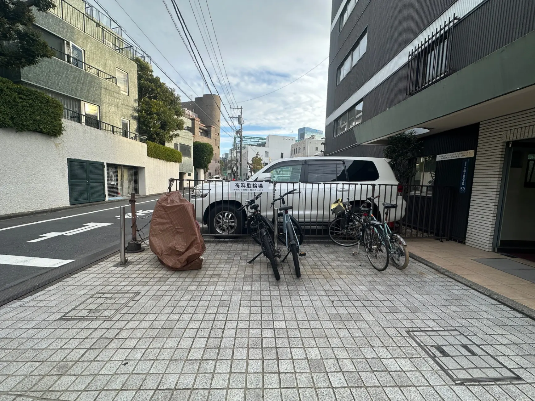 日本の都市部にある舗装された駐車場と自転車置場、複数のアパート建物と植栽、日中の屋外シーン