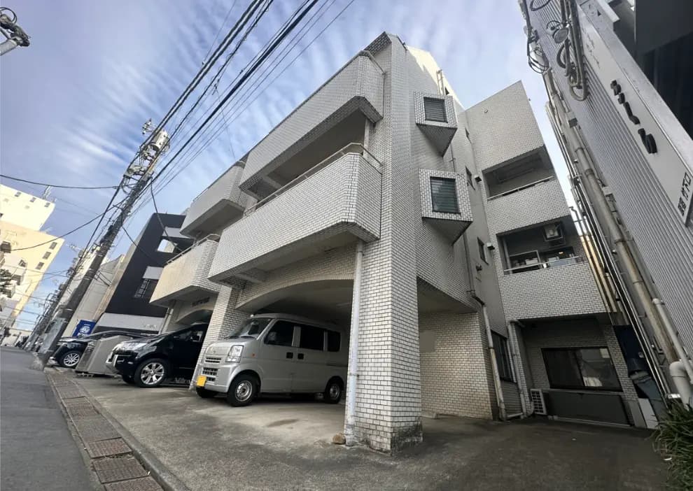 都市の日当たりの良い日に撮影された現代的な建物と駐車スペース、小規模マンション、周囲に電線と他の建物が見える風景