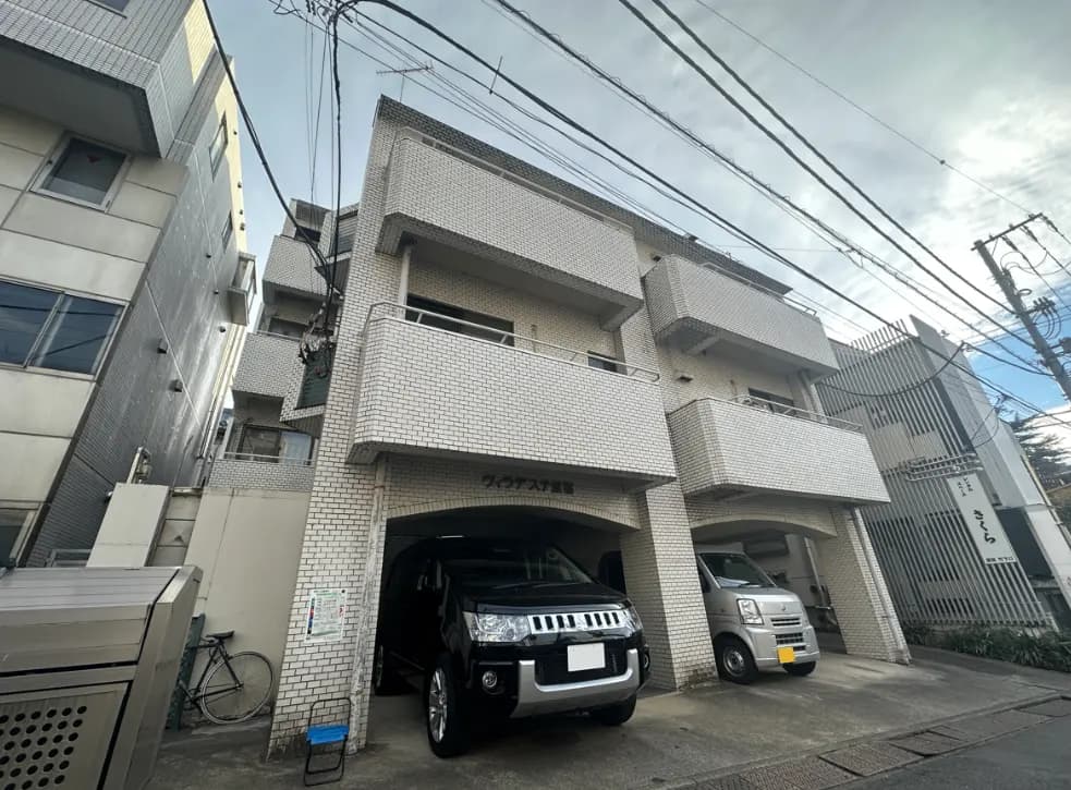 現代的なアパートビルの外観、駐車場に車が2台駐車しており、白く小さなタイルで覆われたファサード